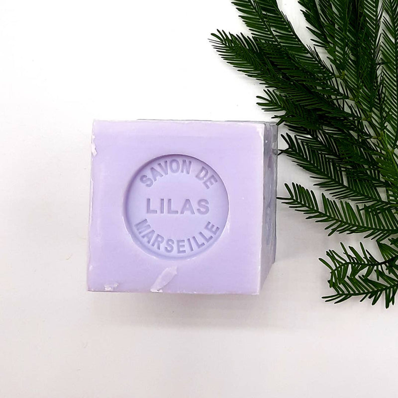 A bar of lilac-colored, paraben-free soap labeled "Senteurs De France Marseille Lilac Cube Soap" rests next to a green fern branch on a white surface.