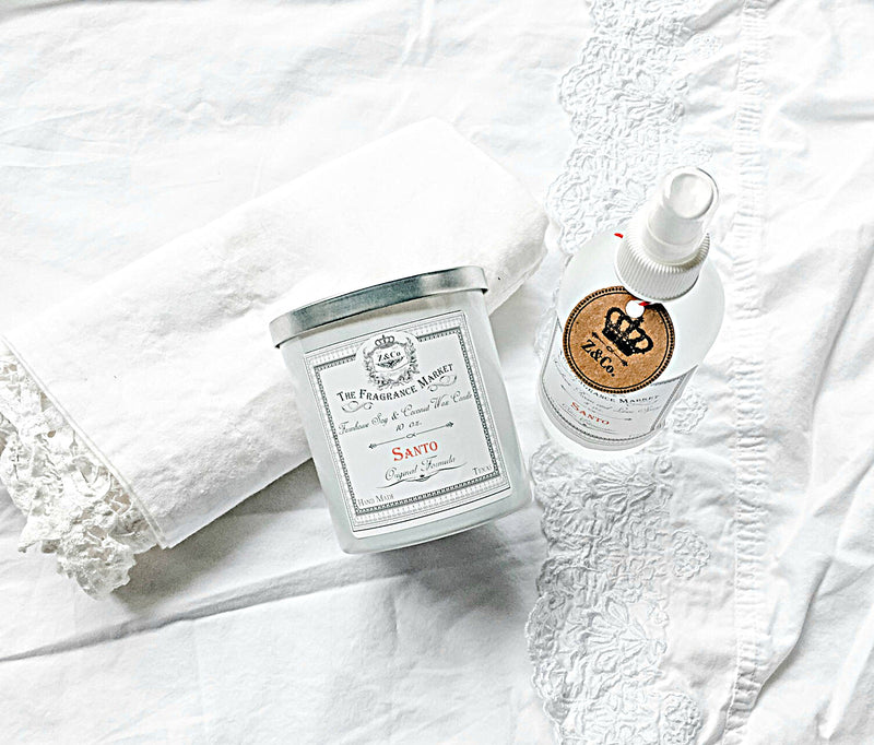Flat lay of skincare products including a scented candle, Z&Co. Santo Farmhouse Room/Linen Spray, and a jar, all on a delicate white lace fabric, conveying a serene and clean aesthetic.