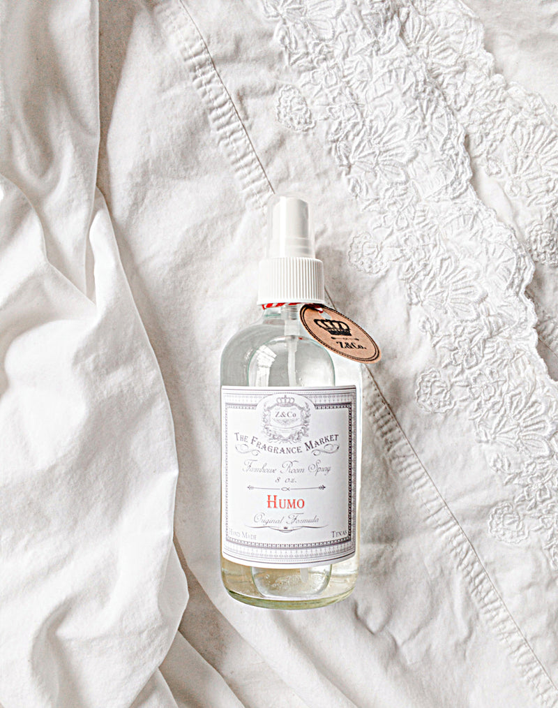 A bottle of Z&Co. Humo Farmhouse Room/Linen Spray, lying on a white textured bedspread. The spray bottle is transparent with a simple label and a white spray head.