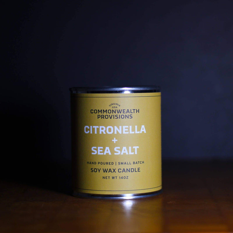 A Commonwealth Provisions Citronella + Sea Salt Candle in a yellow metal tin labeled "commonwealth provisions, citronella + sea salt, hand poured | small batch, soy wax candle, net wt 14oz" on
