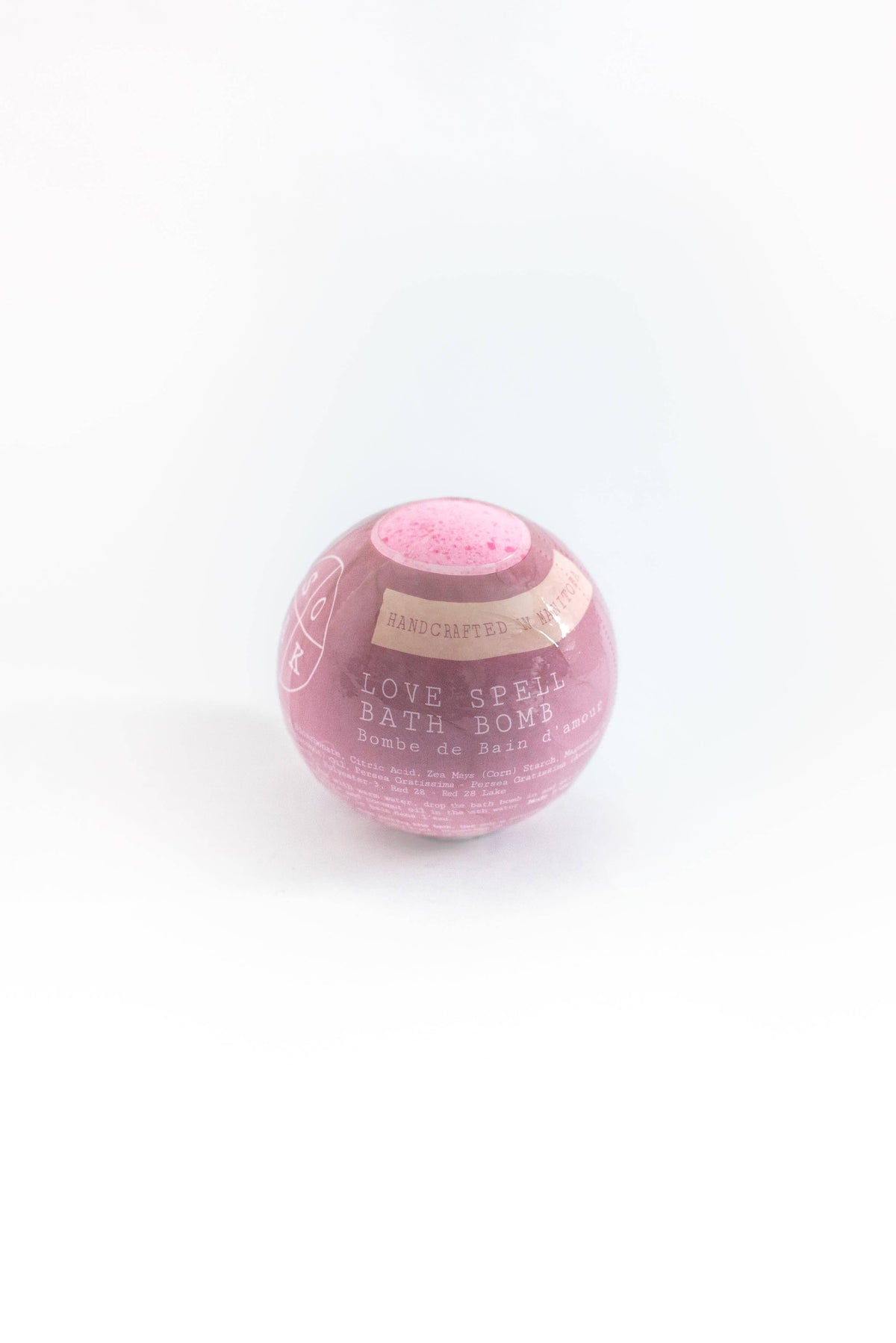 A pink, spherical "SOAK Bath Co. - Love Spell Bath Bomb" displayed against a white background. The packaging is partially transparent shrink wrap and features textual information.
