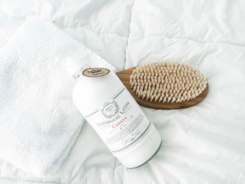 A bottle of Z&Co. Cabana Farmhouse Lotion enriched with Shea Butter beside a wooden brush on a white, crinkled bed sheet, giving a sense of personal care and relaxation.