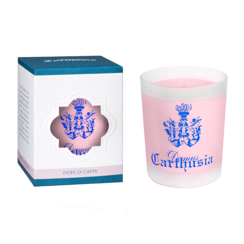 A pink Carthusia Fiori di Capri Candle in a clear engraved glass container beside its packaging box, which is white with a blue top and a heart-shaped cutout revealing a crest design. The text on both reads Carthusia I Profumi de Capri.