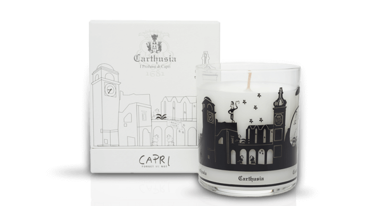 A decorative scented Carthusia Capri Forget Me Not Candle with fig tree essence in a clear glass with black designs, next to its Carthusia I Profumi de Capri white packaging box featuring elegant illustrations and text.
