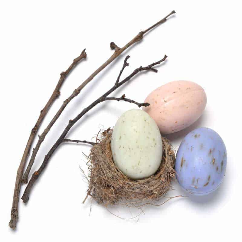 Three colorful La Lavande Egg Carton with 6 Assorted French Egg Soaps in pastel shades of pink, green, and blue resting in a small nest beside some bare twigs, on a white background.