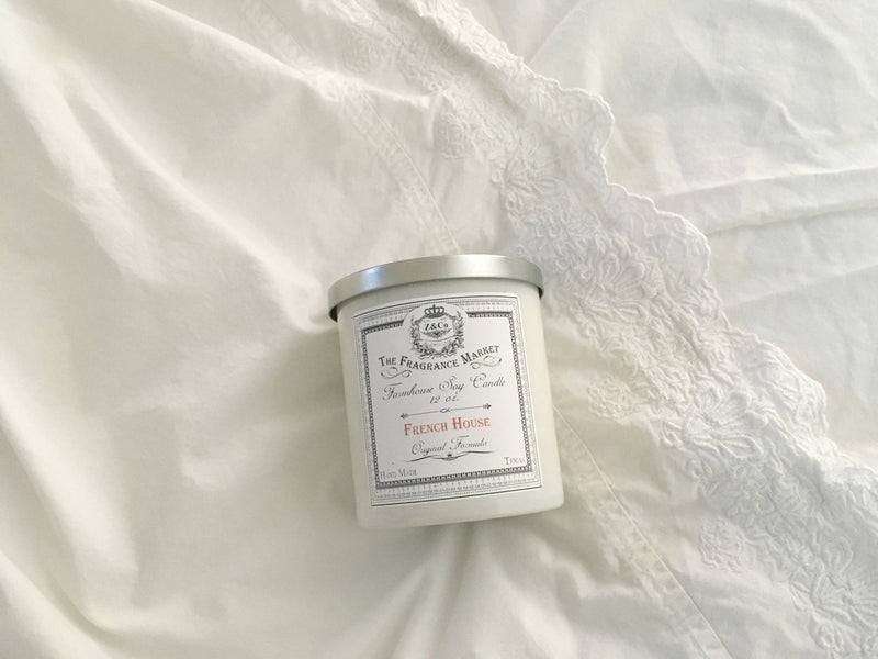 A Z&Co. French House Farmhouse Candle in a tin container, made from soy and coconut wax, placed on a white bedsheet with delicate lace detailing.