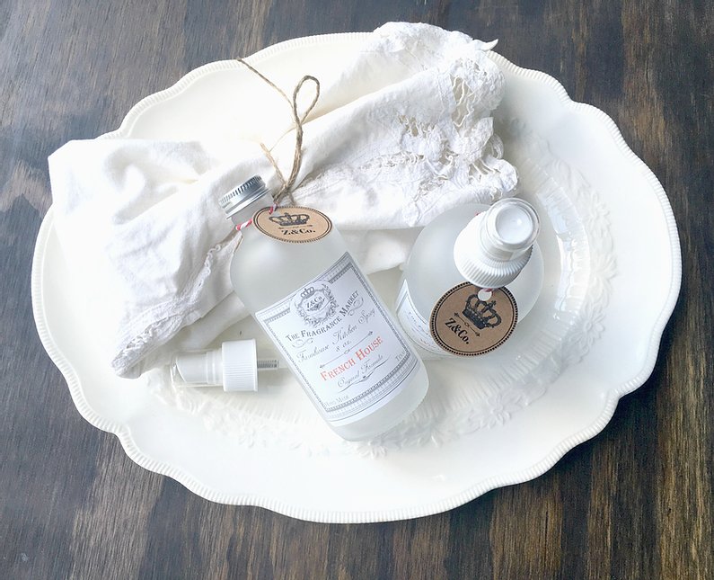 A white tray holding two bottles of lavender hair care products, a Z&Co. French House Farmhouse Room/Linen Spray bottle, and a small piece of natural fabric, all atop a dark wooden surface.