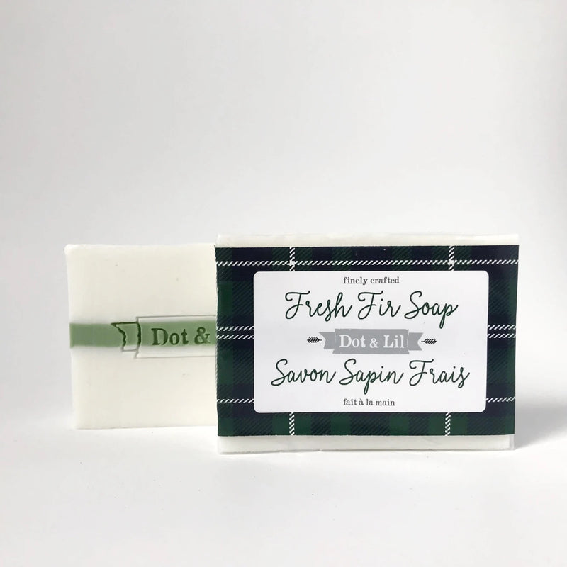 A bar of Dot & Lil Fresh Fir Bar Soap next to its packaging featuring a green and black plaid design and bilingual text. The bar is partially unwrapped on the left side.