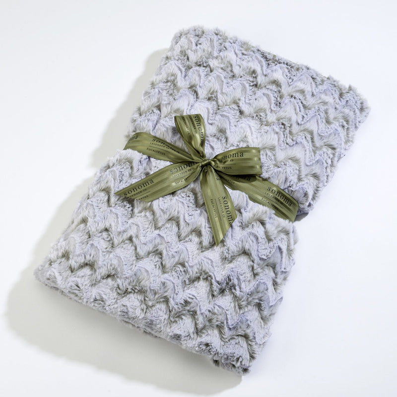 A Sonoma Lavender knitted scarf with a textured pattern, neatly folded and tied with a shiny olive green ribbon featuring gold text, infused with eucalyptus lavender.