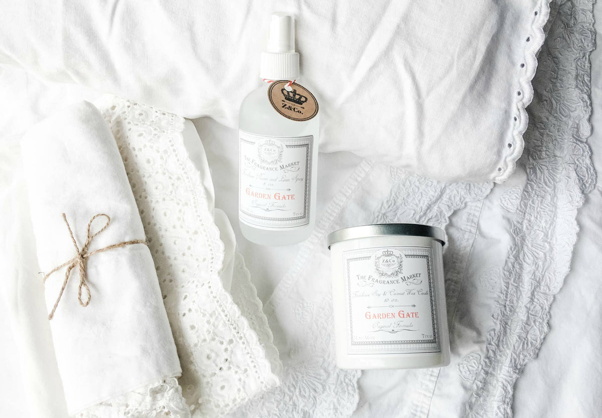 A white bed with a folded lace napkin tied with twine, a spray bottle, and a non-toxic Z&Co. Garden Gate Farmhouse Candle, all arranged in a bright setting with subtle gardenia jasmine fragrance.