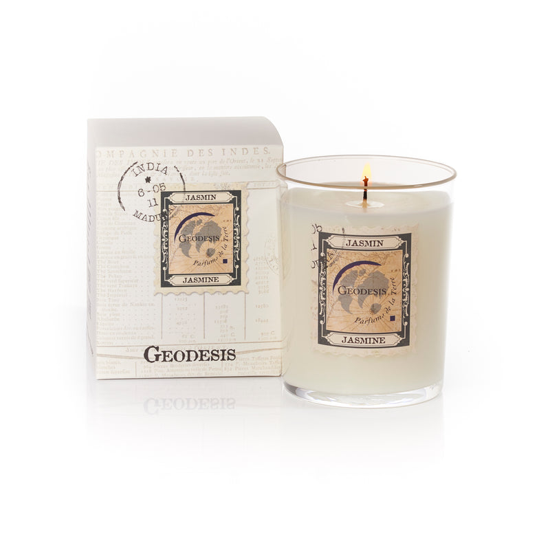 A Geodesis Jasmin 220gm scented candle with a 60-hour burn time in a clear glass holder beside an open book with pages displaying vintage label designs, set against a pure white background.