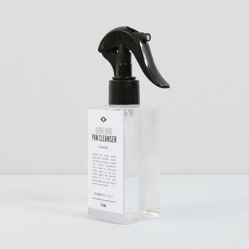 A transparent spray bottle labeled "Norfolk Natural Living Good Dog Coastal Paw Cleanser" with a black spray nozzle, placed on a white surface. The clear liquid is enriched with antibacterial properties, making it ideal for cleaning muddy paws. The label features small text details.