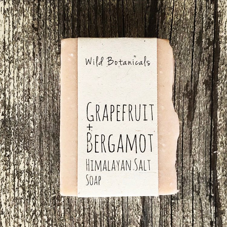 A bar of Wild Botanicals Grapefruit Bergamot Himalayan Salt Soap on a rustic wood background. The soap is packaged in simple, neutral-colored paper with black text.