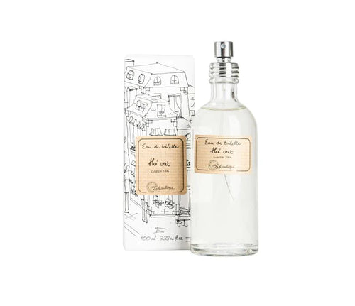 A bottle of Lothantique Green Tea Eau de Toilette with a clear body and a spray nozzle, next to its packaging featuring sketch-like illustrations of a cityscape. The label indicates it is scented with lemon jasmine.