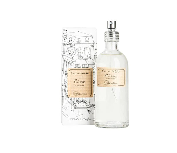 A bottle of Lothantique Green Tea Eau de Toilette with a clear body and a spray nozzle, next to its packaging featuring sketch-like illustrations of a cityscape. The label indicates it is scented with lemon jasmine.