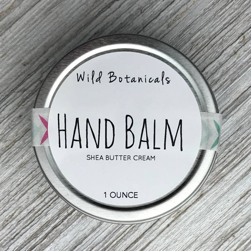 A tin of Wild Botanicals 1oz Hand Balm Tin with organic shea butter cream, labeled clearly on the lid. The container has a white background with simple, modern black and pink text, displayed.