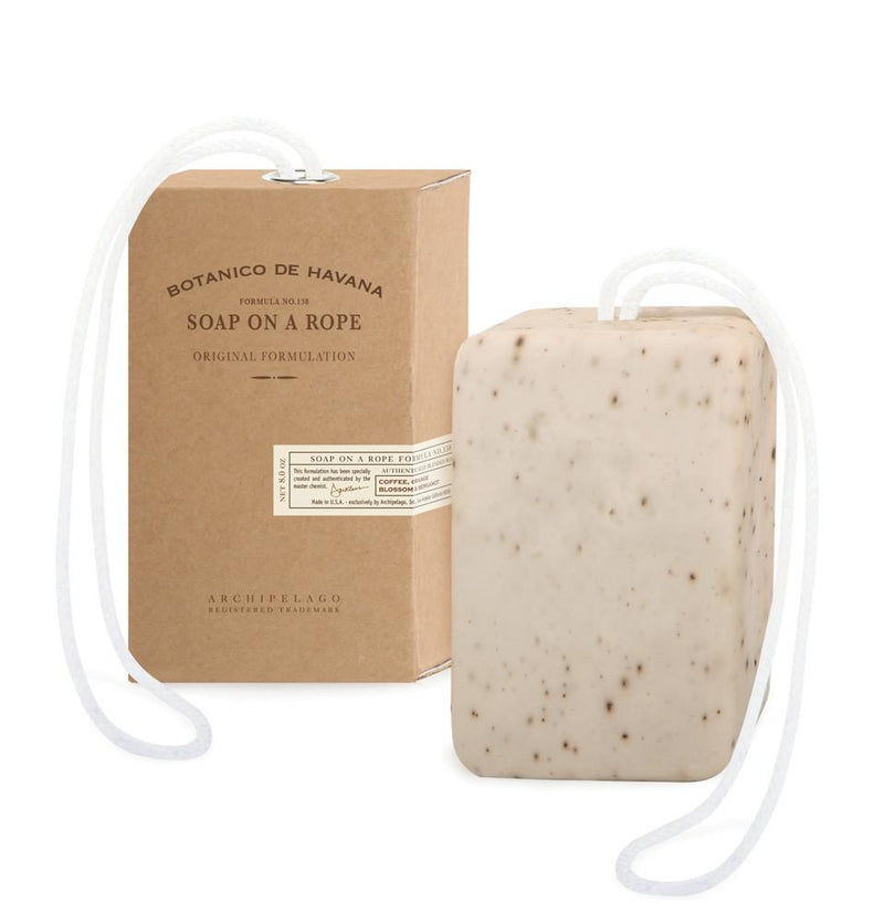 A bar of exfoliating Archipelago Botanico de Havana soap with specks, attached to a white rope, placed next to its brown cardboard packaging labeled "Tobacco Flower Havana Soap on a Rope Original Formulation.