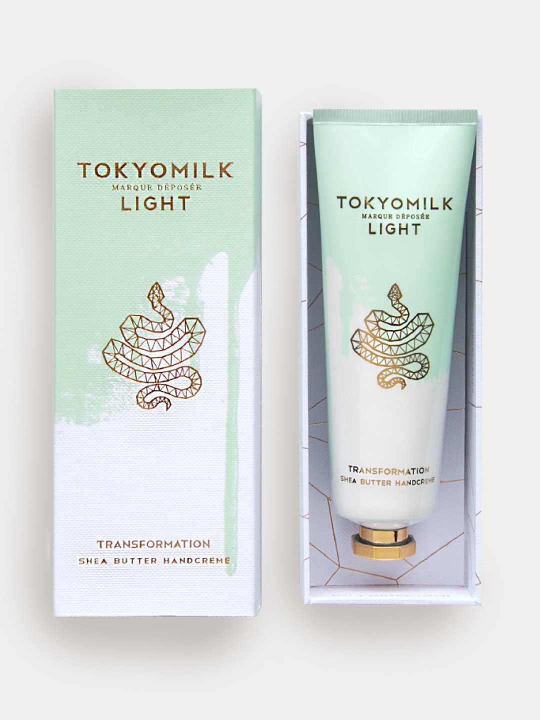 A tube of Margot Elena TokyoMilk Light Transformation No. 03 Shea Butter Handcreme next to its packaging, both decorated with a minimalist white and gold origami boat design on a light green background infused with the scent of