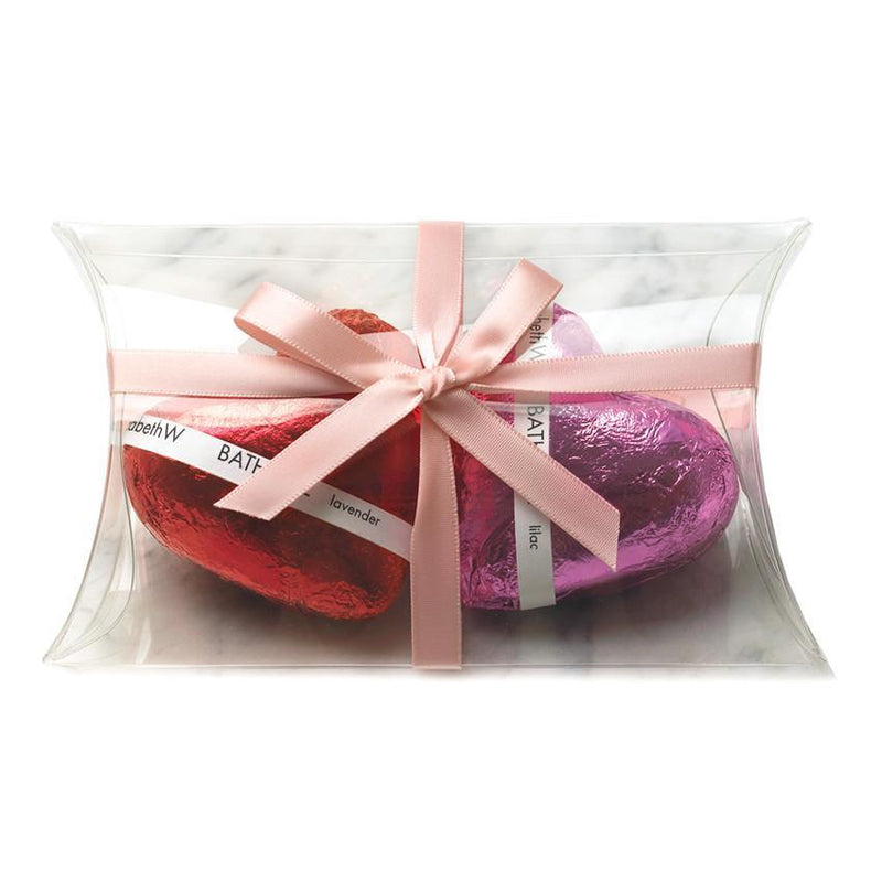 Three colorful, foil-wrapped Easter eggs in red, purple, and pink, transformed into elizabeth W Lavender Fizz Heart bath bombs, are elegantly placed inside a clear, pillow-shaped gift box tied with a soft pink bow.