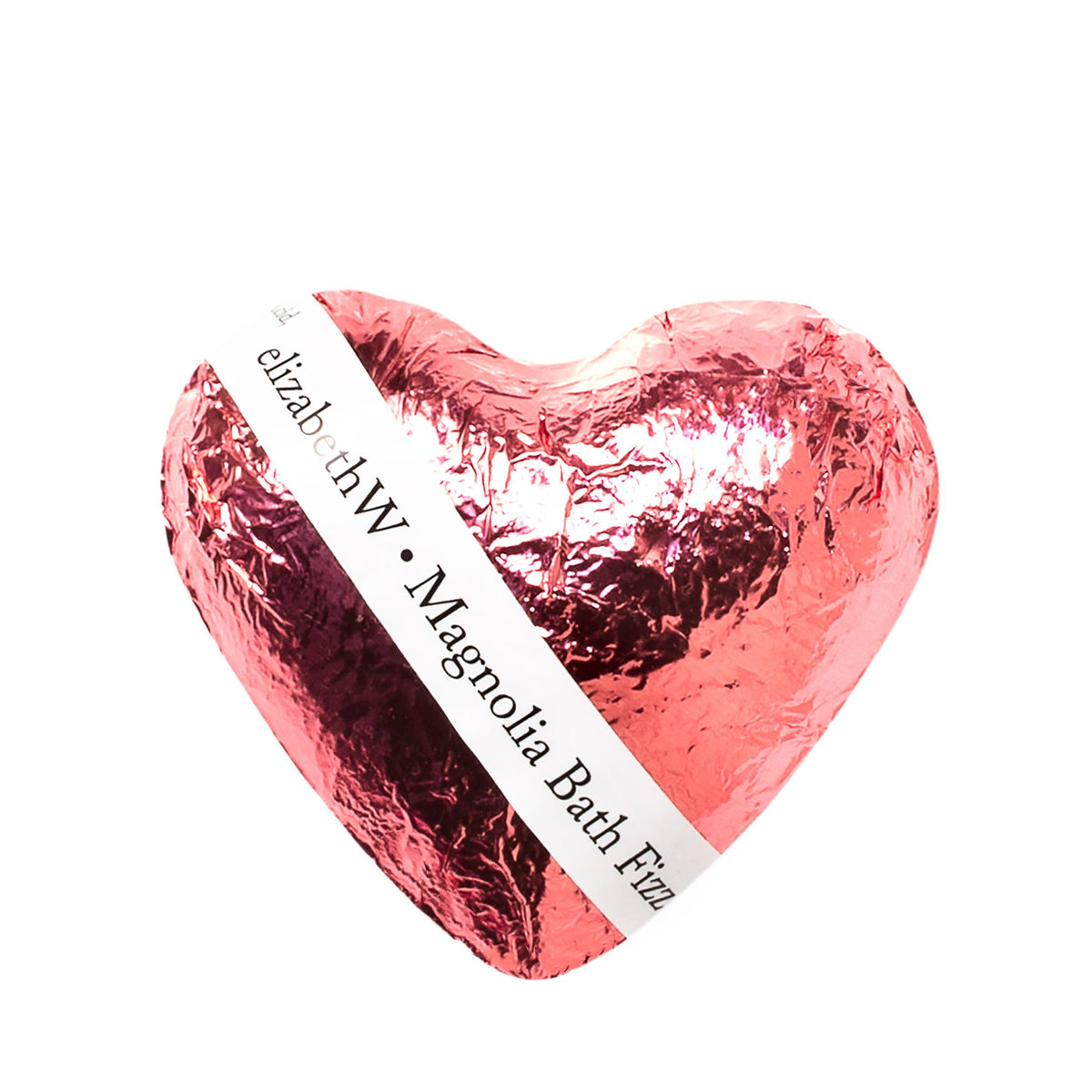A heart-shaped, pink foil-wrapped bath bomb with a label that reads "elizabeth W Fizz Heart - Magnolia." The packaging has a crinkly texture, reflecting studio lighting.