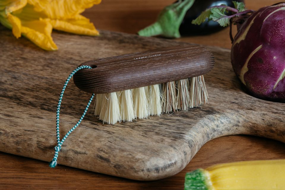 A Andrée Jardin Hard & Soft Vegetable Brush Heritage with stiff bristles and a blue cord lies on a rustic wooden cutting board, surrounded by fresh vegetables including a purple kohlrabi and yellow squash flowers.