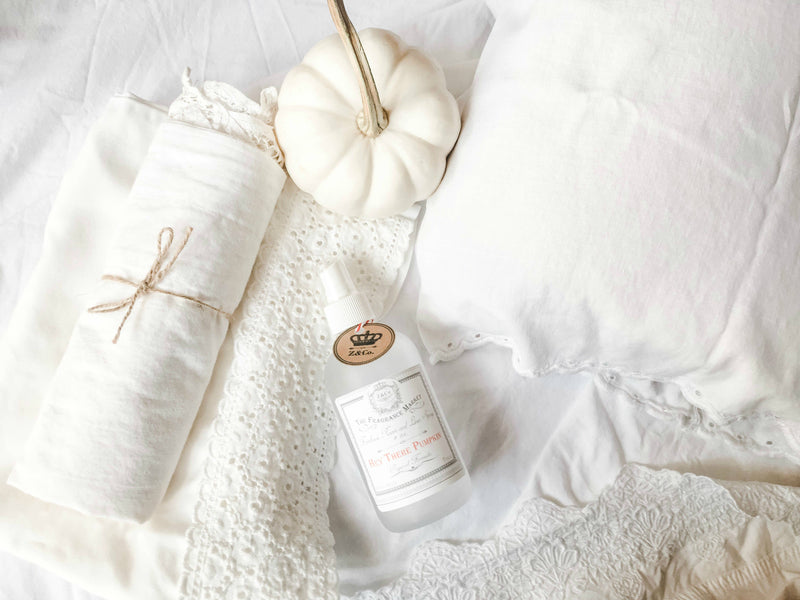 A gentle and serene arrangement on a white bedsheet featuring a white pumpkin, a tied bundle of linen, and a bottle of Z&Co. Hey There Pumpkin Farmhouse Room/Linen Spray, all surrounded by delicate lace textures.