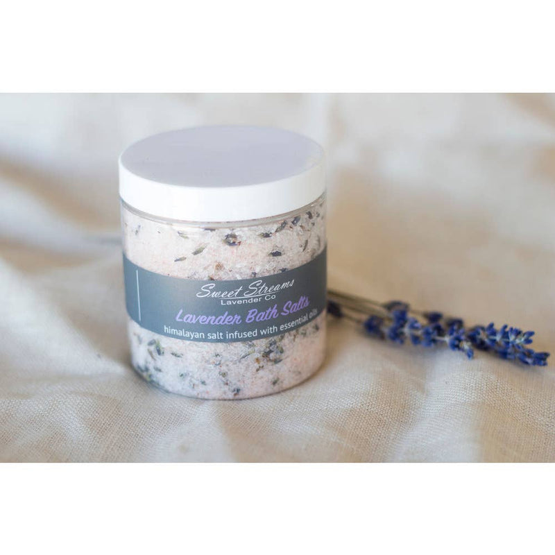 A jar of Sweet Streams Lavender Co. - Lavender Bath Salts labeled "sweet stream Himalayan lavender bath salts" with dried lavender sprigs beside it, set on a textured beige fabric.