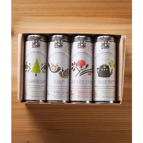 A Flying Bird Botanicals - Holiday Cheer Gift Box containing five cylindrical tea tins with elegant, botanical-themed labels, arranged neatly on a wooden surface. Each tin displays unique floral and herb illustrations, including Vanilla Rose Ceylon and Cedarwood.