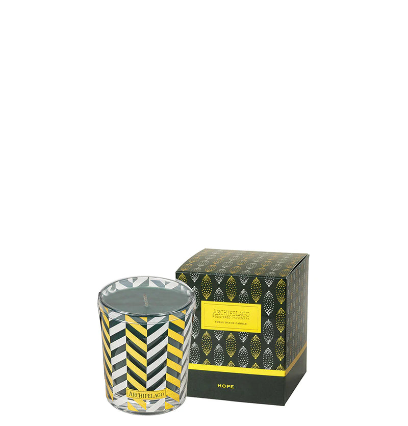 A decorative Archipelago Botanicals Hope Boxed Votive Candle in a clear glass jar next to its packaging box. The box and candle have a geometric pattern in black, gold, and green with the word "hope" printed on them.