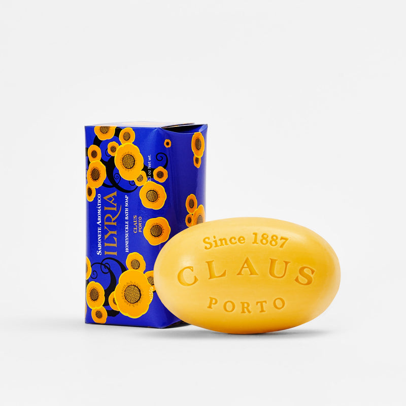 A yellow Claus Porto Ilyria Honeysuckle soap bar beside its blue packaging decorated with orange and yellow flowers. The soap and packaging feature the brand name "Claus Porto 1887" since 1887.