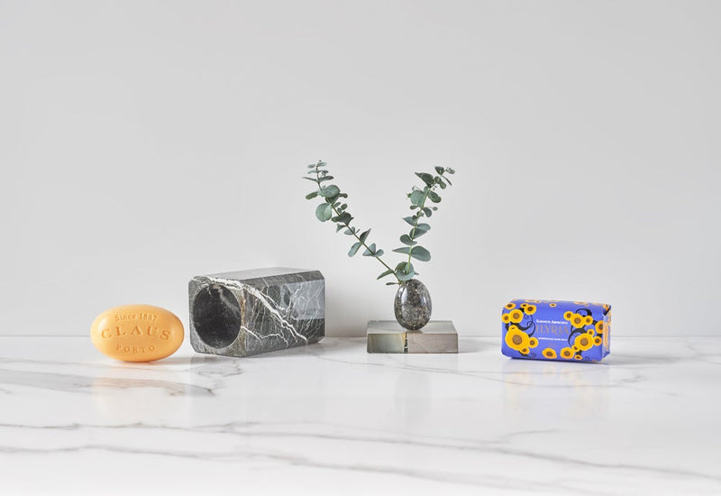 A minimalist display featuring three objects on a marble countertop: a round soap labeled "Claus Porto Ilyria Honeysuckle Soap - 150gm," a marble cylinder with a eucalyptus branch, and a colorful pattern.