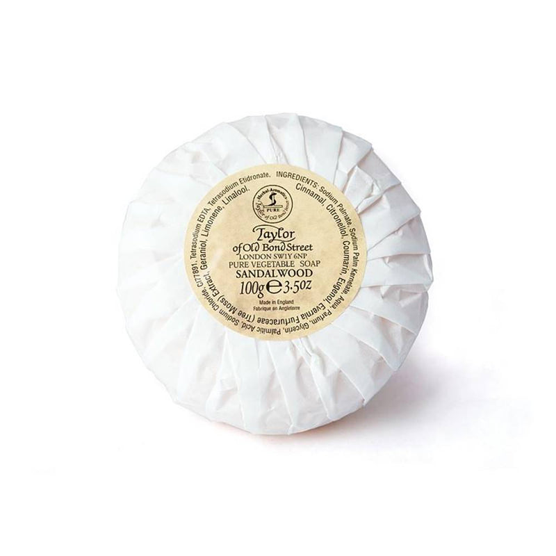 A round bar of Taylor of Old Bond Street Sandalwood Hand Soap - 100gm, wrapped in white pleated paper with a gold and black label on top, suitable for sensitive skin.