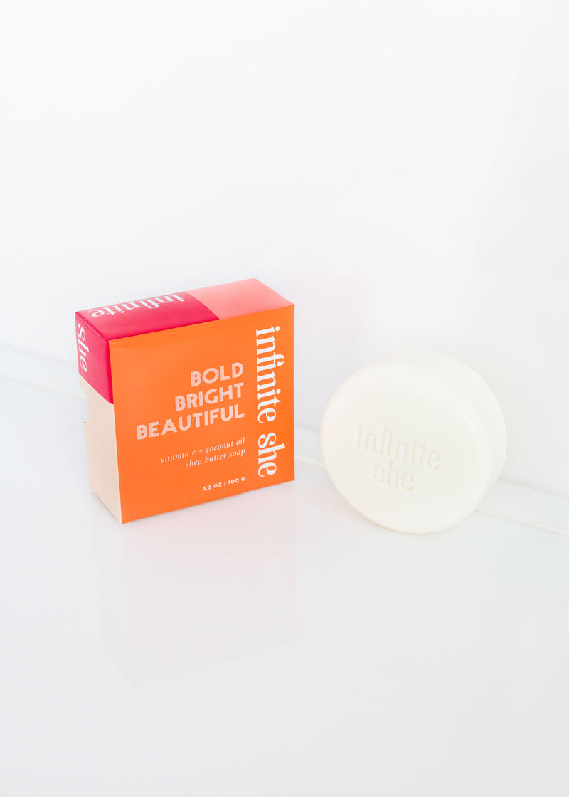 A bright orange and pink Margot Elena product box labeled "bold bright beautiful" next to a white compact with "Infinite Shea" written on the top, set against a white background.