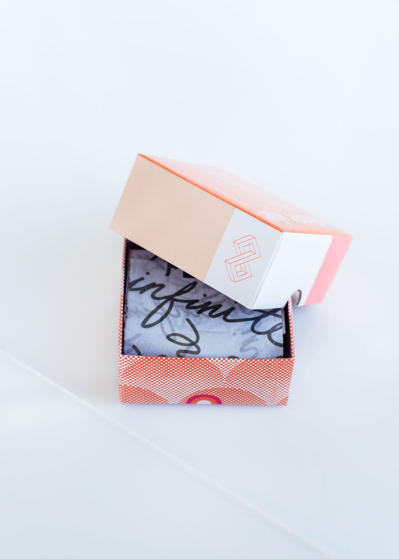 An open gift box with a light pink lid slightly askew, revealing Margot Elena's Infinite She Bold Bright Beautiful Shea Butter soap with a polka dot pattern inside, against a white background.