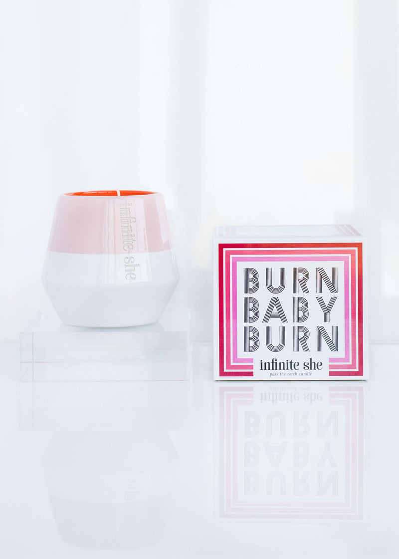 A stylish Infinite She Burn Baby Burn Ceramic Candle by Margot Elena, next to a sleek, white and peach-colored container with hints of Fresh Cut Iris, set against a bright, reflective surface with a soft