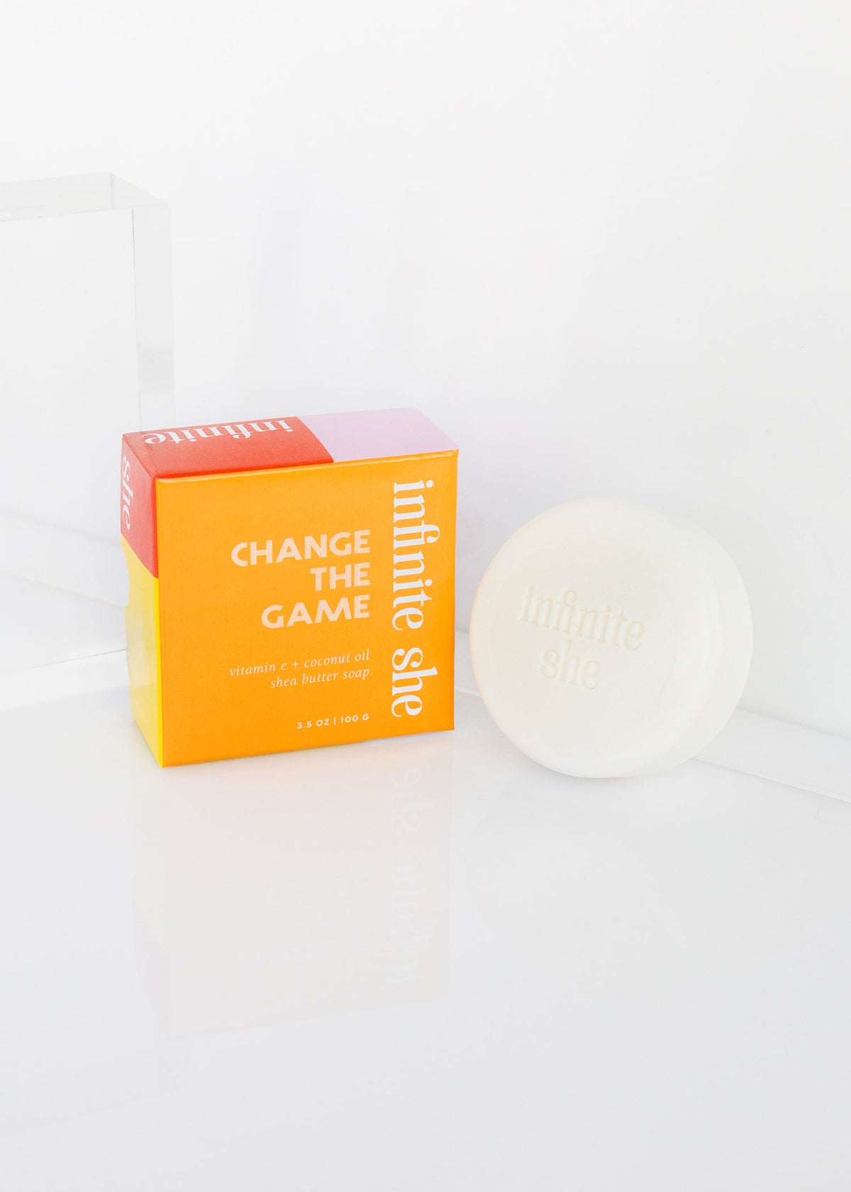 A skincare product packaging labeled "Infinite She Change the Game Shea Butter Soap" next to a round container with "Infinite She" on the lid, set against a clean, white backdrop. The product contains Coconut Oil.