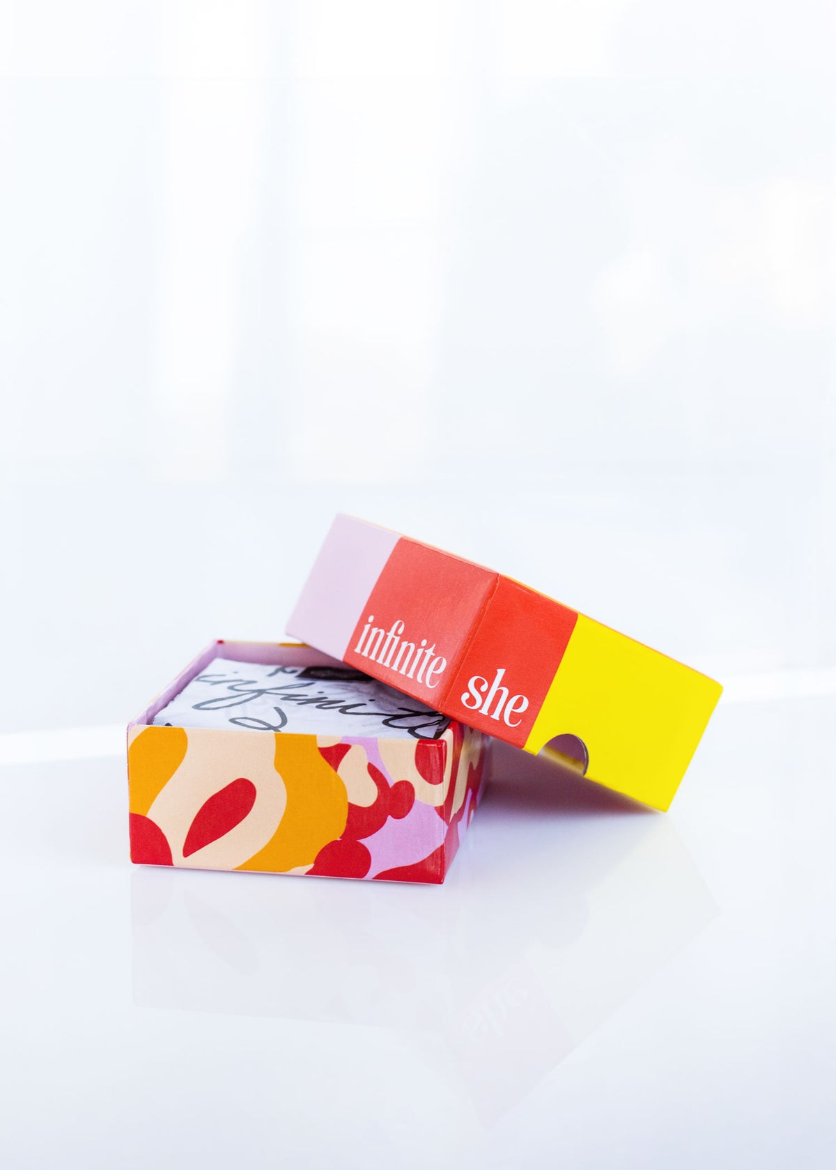 A colorful Margot Elena product box with the words "Infinite She Change the Game Shea Butter Soap" on a vibrant red label, partially opened to reveal a Shea Butter soap patterned interior, set against a bright, light background.