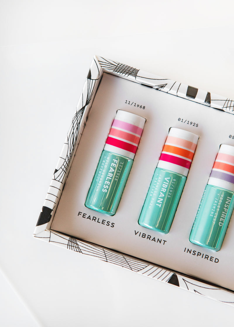 Three colorful lip balms labeled "fearless," "vibrant," and "inspired" inside an open Margot Elena Infinite She Essence Exploration Eau de Parfum Discovery Set against a white background.