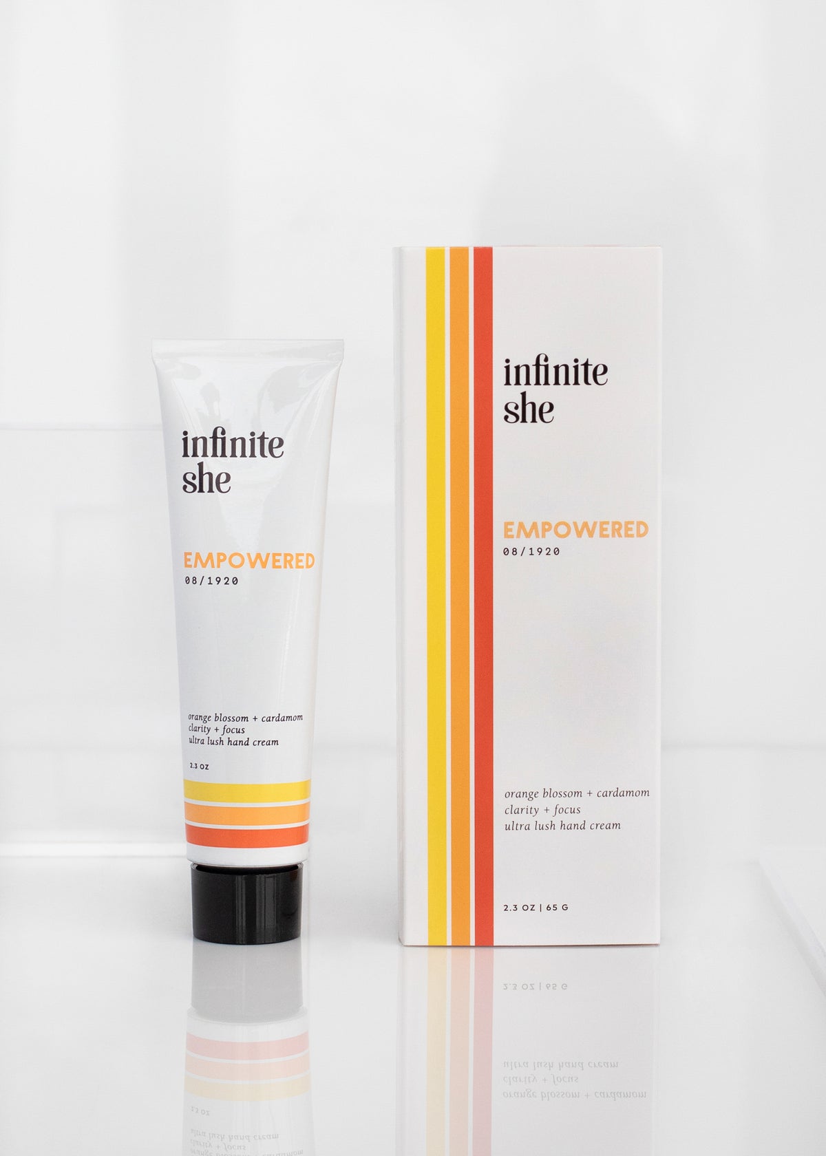 A tube of Margot Elena's Infinite She Empowered Ultra Lush Hand Cream next to its packaging. The items are labeled "empowered" and display an orange, yellow, and white color theme on a white background.