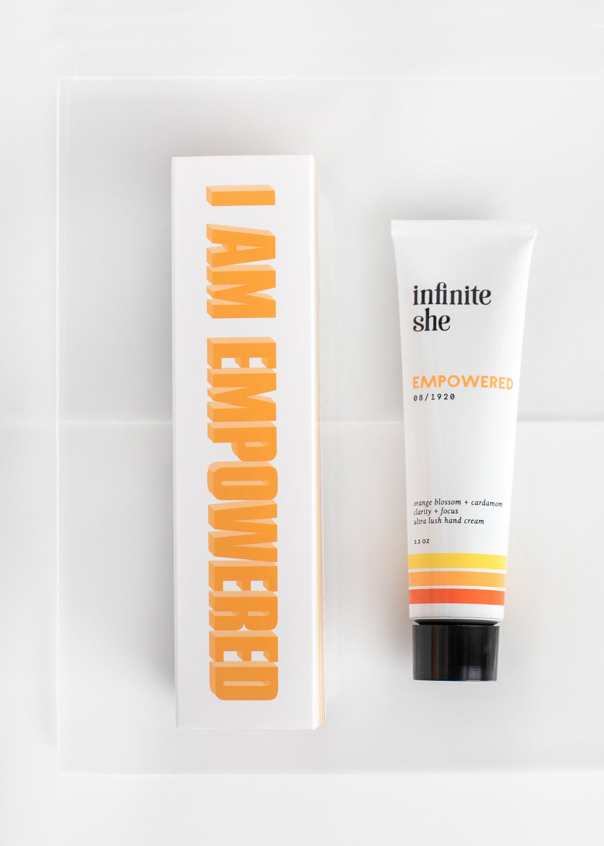 Orange and white packaging marked "i am empowered" alongside a tube of vegan hand cream labeled "Infinite She Empowered Ultra Lush Hand Cream" by Margot Elena on a clean, bright background.