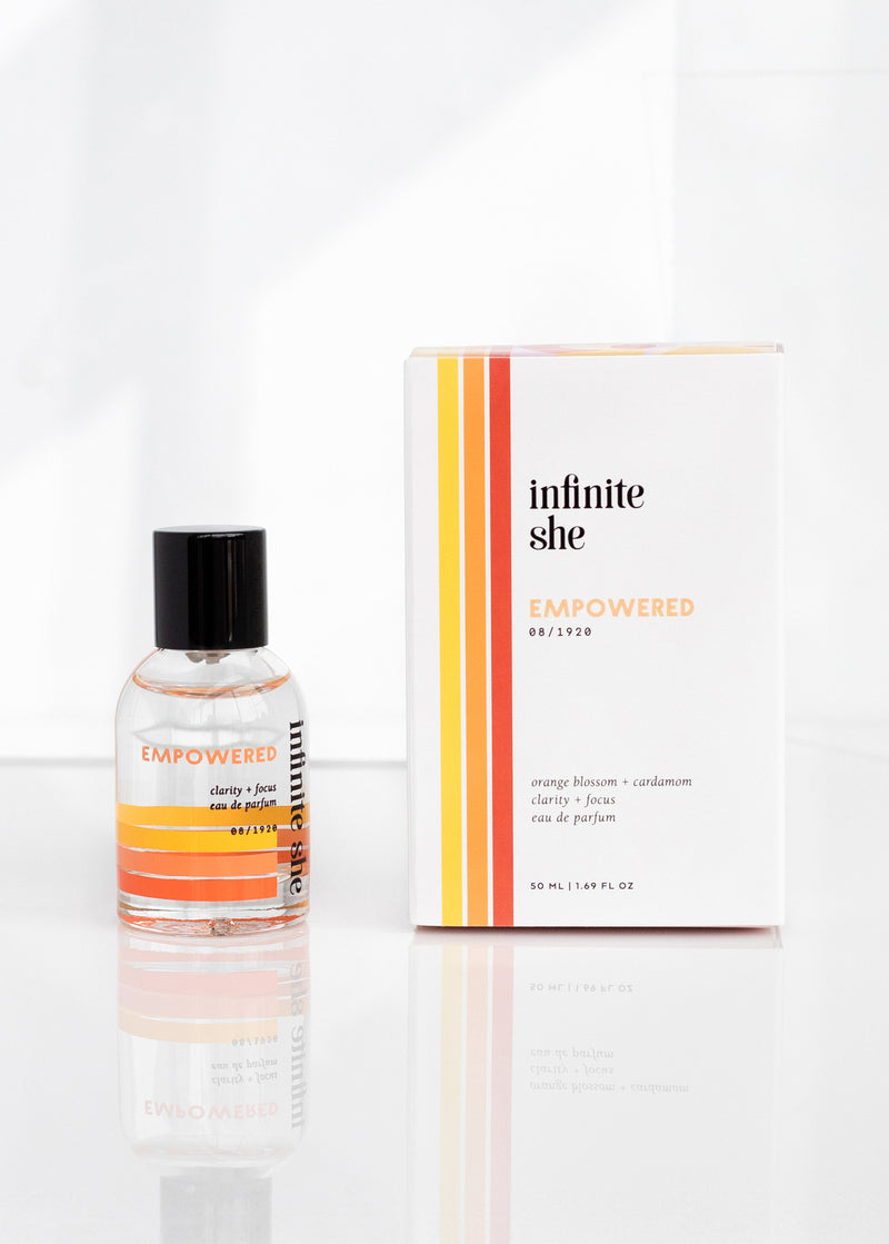 A bottle of Margot Elena's "Infinite She Empowered Eau de Parfum" with orange blossom and cardamom scents, accompanied by its white packaging, standing on a reflective surface with a white background.