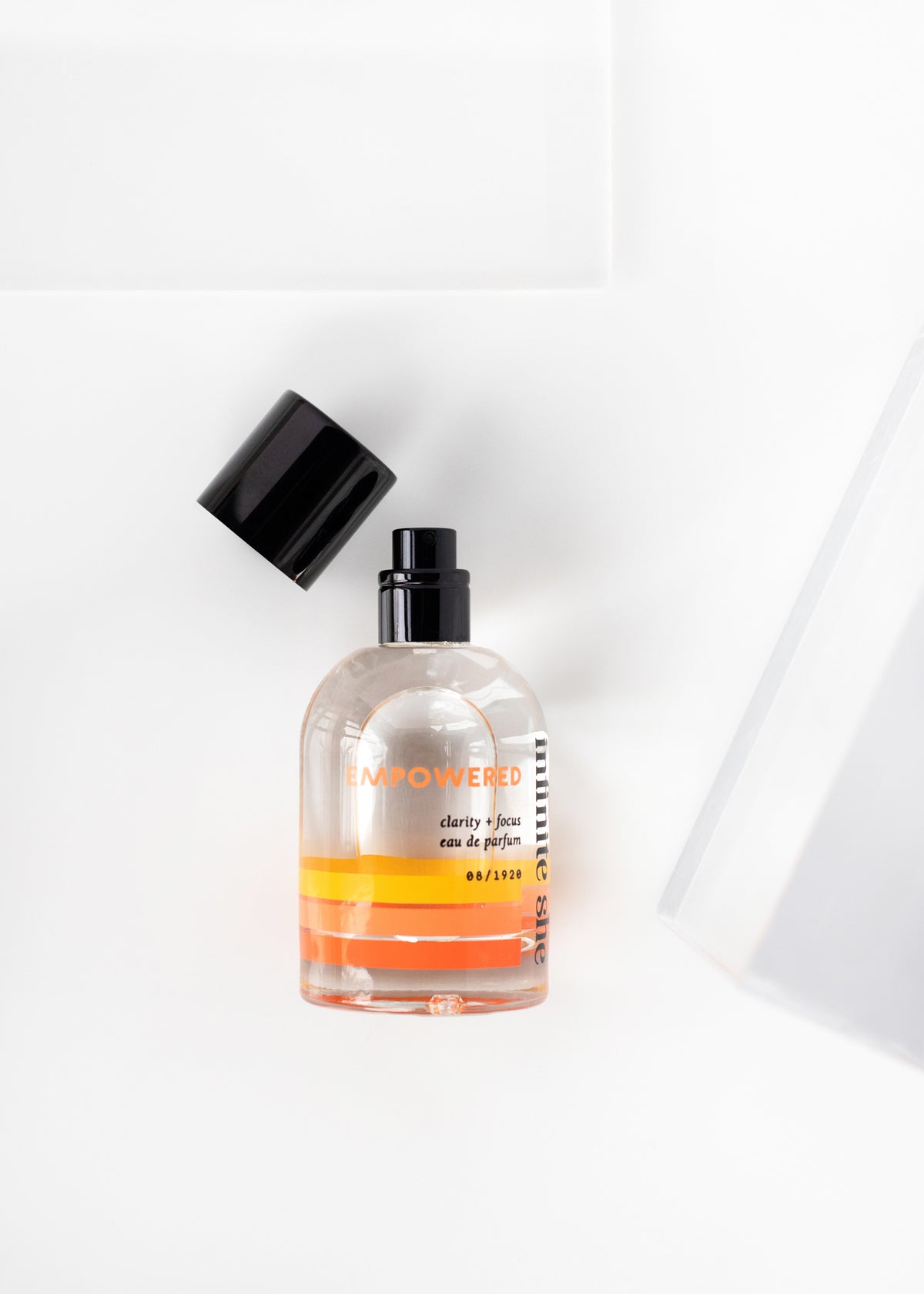 Transparent spray bottle labeled "Infinite She Empowered Eau de Parfum" with orange blossom liquid and a black cap, set on a white surface with a soft shadow from Margot Elena.