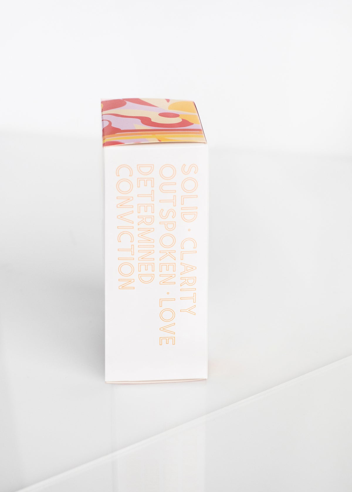 A slim box stands on a bright white surface, displaying Margot Elena's Infinite She Empowered Eau de Parfum in black text, complemented by colorful abstract art with hints of orange blossom.