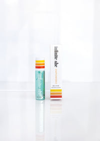 Two Margot Elena beauty products, a small tube and a larger box with modern design, labeled "Infinite She Empowered Rollerball Eau de Parfum," displayed against a clean, bright white background. Both items are infused with the vegan fragrance of orange.
