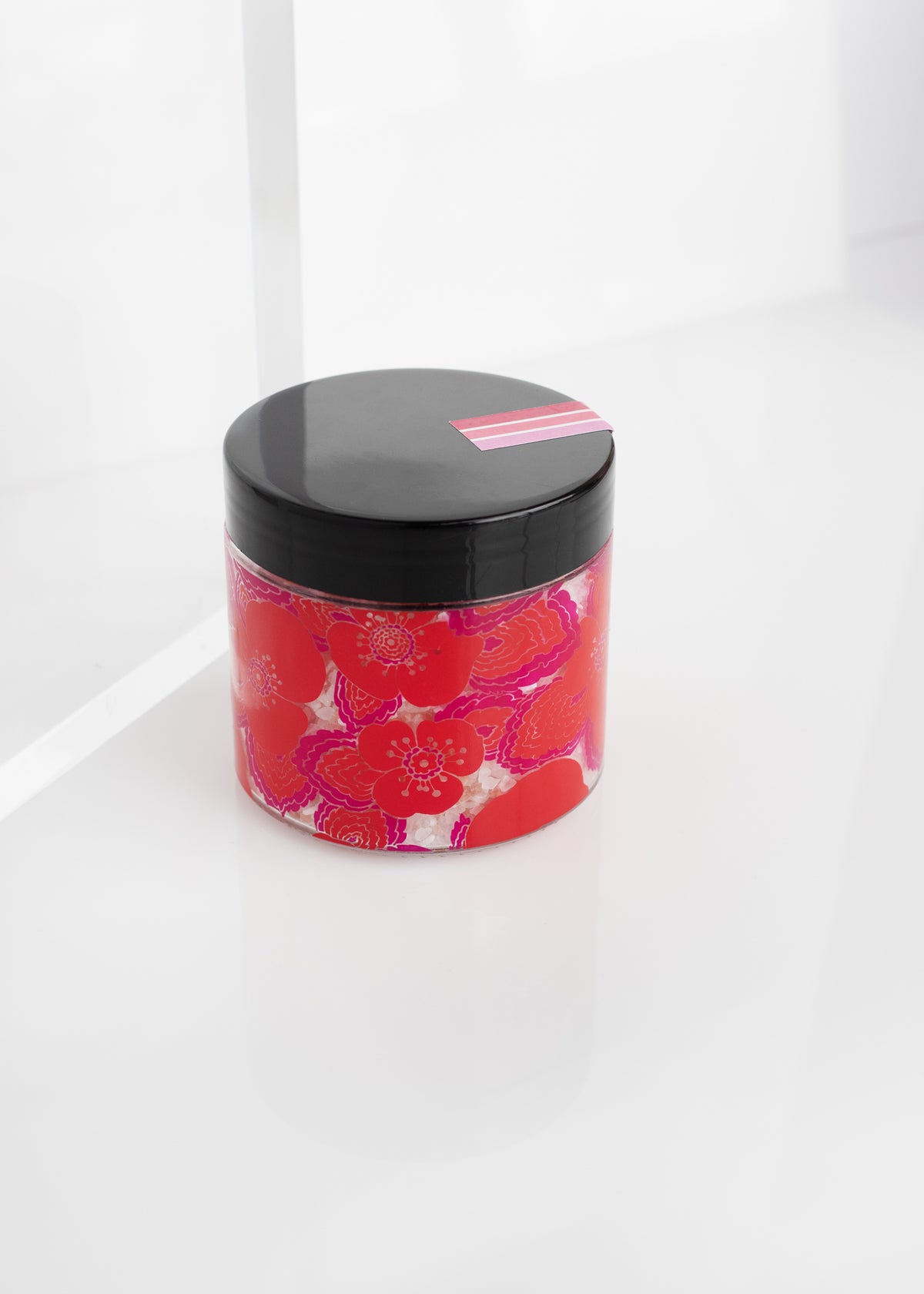 A Margot Elena cosmetic jar with a black lid and a Wild Geranium + Rosewood pattern on the body, placed on a white reflective surface in a bright interior.