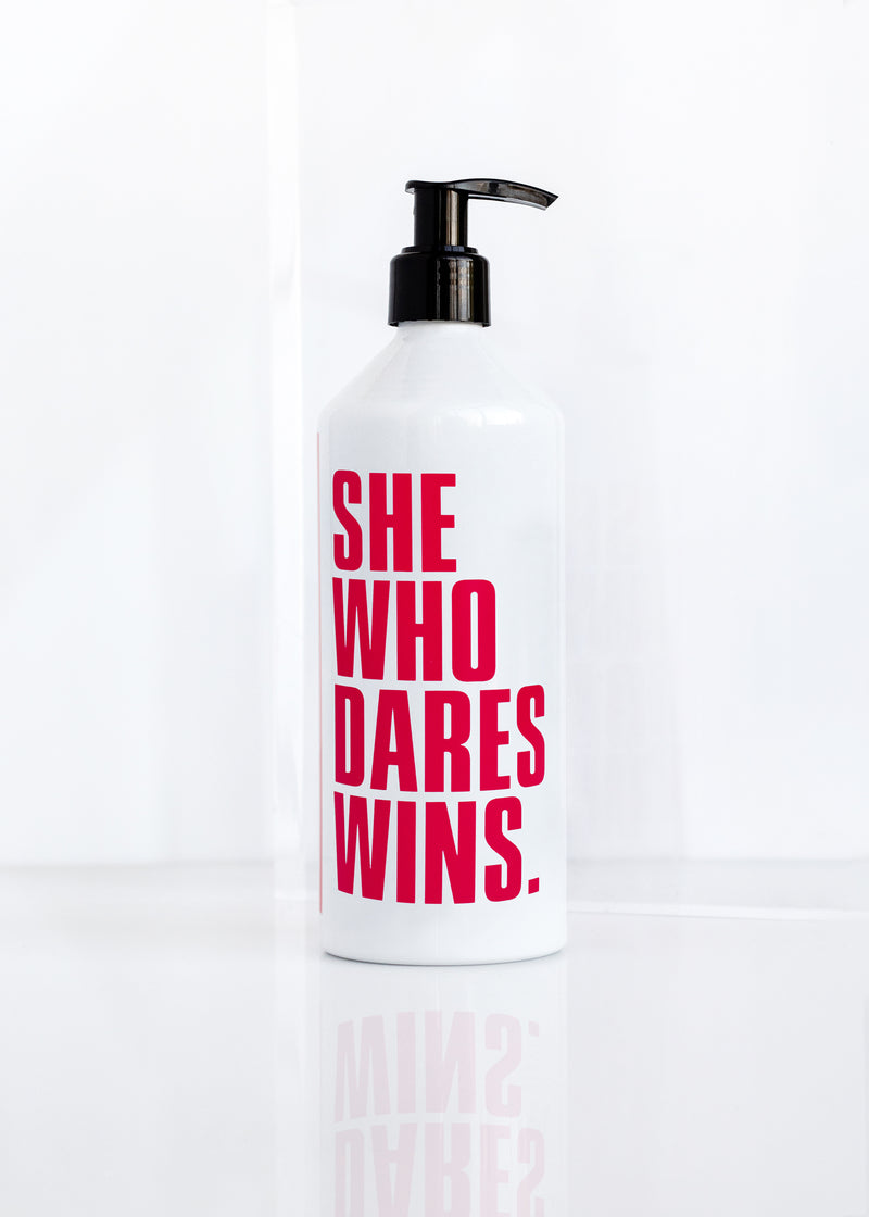 A white bottle of Margot Elena's Infinite She Fearless Hydrating Body Lotion with a pump dispenser and the phrase "she who dares wins" printed in bold pink letters. The bottle is set against a rosewood background.