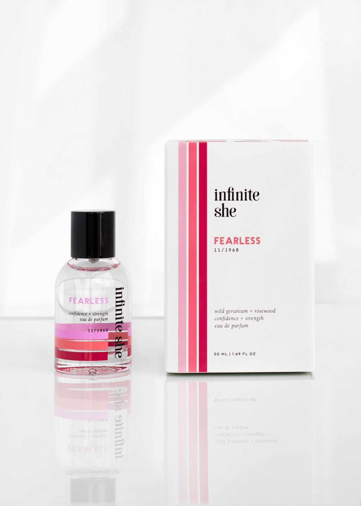 A bottle of Margot Elena's Infinite She Fearless Eau de Parfum next to its packaging, on a reflective surface with a white background. The package features pink stripes and rosewood fragrance notes.