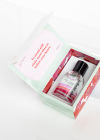 An open Margot Elena beauty subscription box containing a variety of skincare and makeup products, all infused with vegan fragrance, with a motivational card visible on top. The box design features a pink and white color scheme.