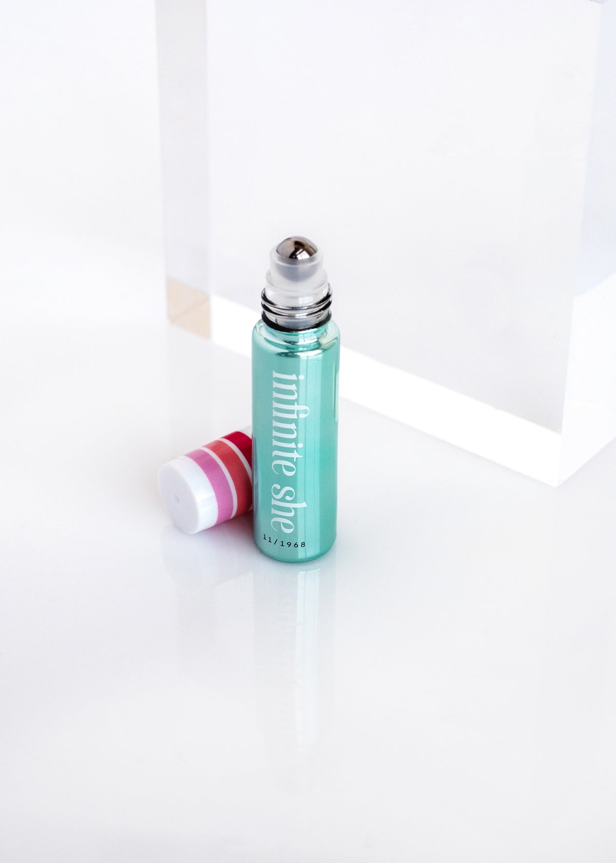 A teal rollerball perfume bottle labeled "Infinite She Fearless Rollerball Eau de Parfum" by Margot Elena, next to its pink cap, displayed against a clean white background with a subtle shadow.