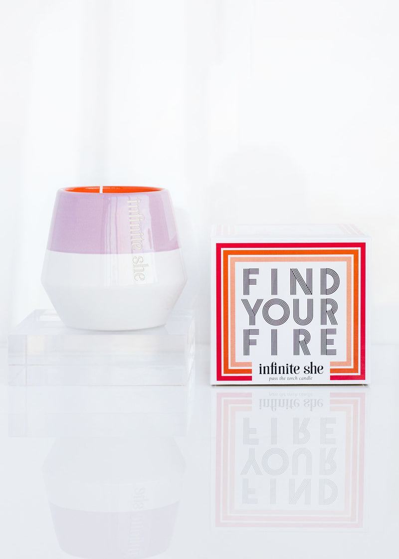 A white and purple candle branded "Infinite She" next to a red and white box with the inscription "Find Your Fire" on a reflective surface, infused with the scent of honeyed amber.